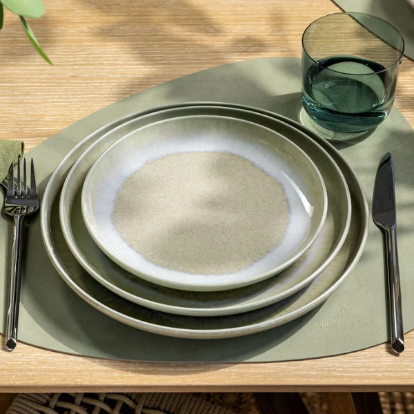 Vista dall’alto di un coperto con piatti verde tenue Perlemor Alga di Villeroy & Boch, disposti a strati in diverse tonalità, abbinati a un bicchiere d’acqua Like Sage. L’insieme riflette un’estetica naturale, calma e terrosa, con carattere artigianale e sottili variazioni cromatiche.