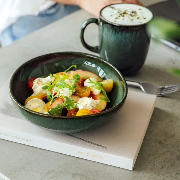 A salad with tomatoes, lettuce and cheese is presented on a book in a green Lave bowl from Villeroy & Boch, accompanied by a frothy drink in a matching green mug on a grey background.
