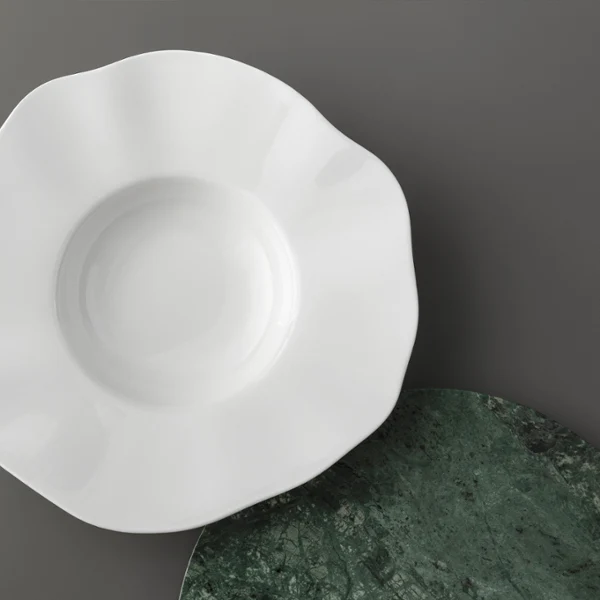 Une assiette Pura blanche à bord festonné est placée sur une surface grise à côté d'un tapis rond vert texturé.