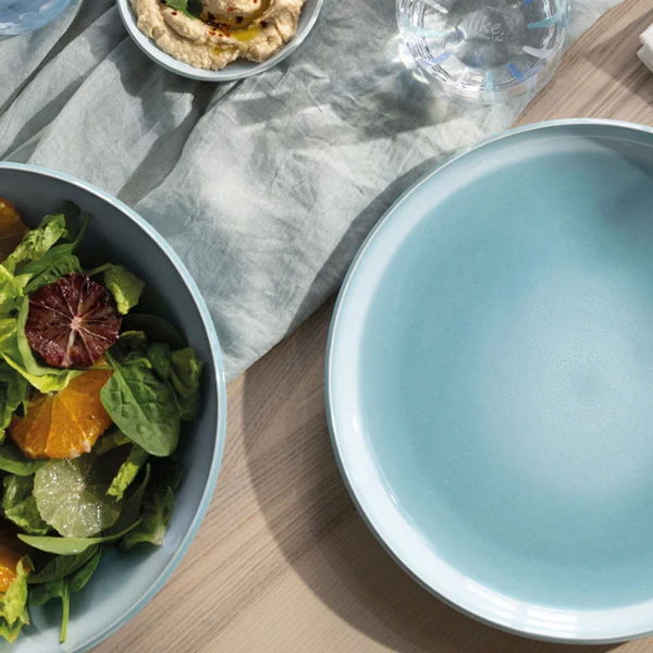 Un piatto vuoto blu madreperla Aqua è posizionato su un tavolo di legno accanto a una ciotola di insalata, posate e un tovagliolo.