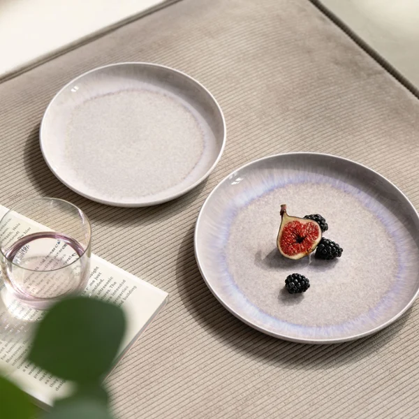 Vista ravvicinata di una tavola con piatti beige tenue Perlemor Sand di Villeroy & Boch e un bicchiere d’acqua Like Pearl. Un piatto è decorato con un fico tagliato a metà, che aggiunge un accento naturale e sottile alla disposizione elegante. La scena mette in risalto le texture organiche e un design calmo e armonioso.