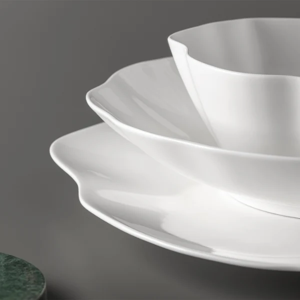 Trois assiettes blanches Pura à bord dentelé sont empilées sur une surface grise à côté d'un objet rond vert.