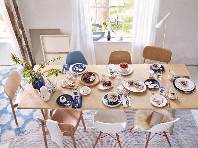 Old Luxembourg dining table set with blue and white dishes, various desserts, teacups, and a small plant. Assorted chairs surround the table, and a window shows a garden view.