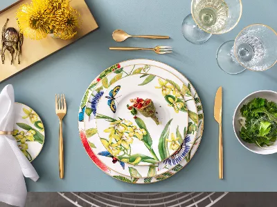 Amazonia - Floral details and golden highlights Plate with tropical flower motif and gold accents on an elegant table, surrounded by flatware and glasses.