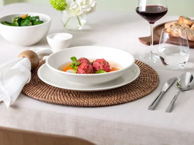 Une table dressée avec une assiette Artesano contenant des boulettes de betterave, des carottes et un peu de verdure. Un verre de vin rouge, de l'eau, un saladier, du pain et des couverts sont disposés sur un set de table tissé et une nappe blanche.