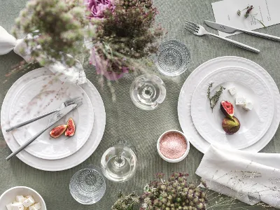 Cellini dinnerware for 2 people on a set table Cellini plates from Villeroy & Boch with figs, cheese, and herbs are set on a beautifully set table. Glasses, flatware, delicate flower arrangements, and a bowl of salt on a green tablecloth complete the ambience.