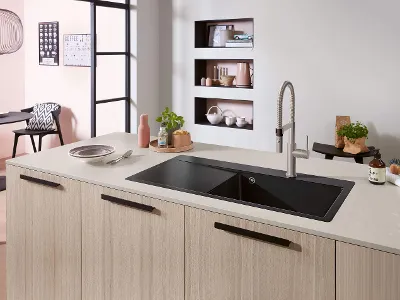 Lavello Subway Style in cucina ad isola chiara con miscelatore Una cucina moderna con un lavello nero su un'isola in legno chiaro, un rubinetto, una pianta e vari oggetti.