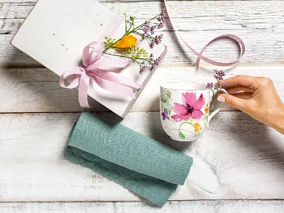 A hand reaches for a Villeroy & Boch Mariefleur mug next to a gift box with a pink ribbon and flowers, placed on a wooden surface with a teal cloth.