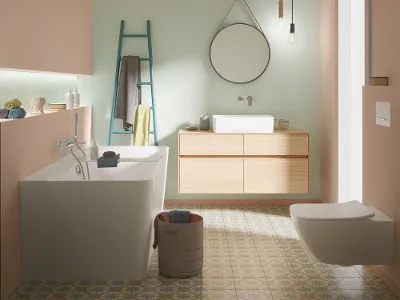 WC e piano lavabo Collaro in bianco, abbinati a un mobile sottolavabo in rovere in un bagno dal design moderno. Bagno moderno con WC Collaro, piano lavabo bianco e mobile sottolavabo in rovere con specchio rotondo. Il pavimento è realizzato con piastrelle a fantasia e il soffitto è disegnato con pannelli di legno.