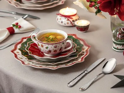 Una tavola festiva con una tazza di zuppa, piatti impilati, posate, candele e decorazioni natalizie.