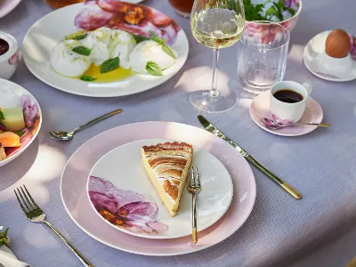 Ein gedeckter Tisch zum Brunch mit Rose Garden Geschirr Elegantes Brunch-Ambiente mit Geschirr aus der Seire Rose Garden, einem Stück Kuchen auf einem Rose Garden Teller, frischem Obstsalat, Kaffee und einem Glas Weißwein.