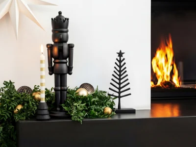 A black nutcracker, a candle and a metal tree Un cascanueces negro, una vela y un árbol de metal se encuentran sobre una repisa de chimenea decorada, junto a una chimenea encendida.