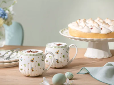 Two Easter Delight mugs on a wooden table Two Easter Delight mugs and a lemon meringue pie on a cake stand sit on a wooden table with plates, cutlery, and a napkin.