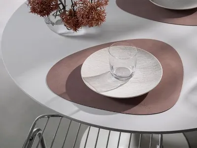 Lifestyle Accessoires con Manufacture Un bicchiere e un piatto poggiano su una tovaglietta astratta marrone di Villeroy & Boch Lifestyle Accessories su un tavolo bianco, con una sedia di metallo e un vaso di fiori secchi sullo sfondo.