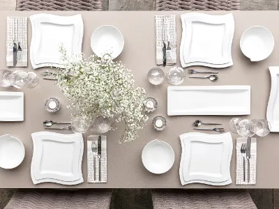 Tavolo per 4 persone apparecchiato con stoviglie NewWave Il set da tavola NewWave di Villeroy & Boch comprende piatti quadrati bianchi, ciotole, posate, bicchieri e un centrotavola con fiori bianchi su una tovaglia beige per quattro persone.
