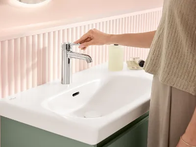 En kvinde står foran den hvide vask og betjener vaskarmaturet i farven krom - begge produkter fra Skyla badeværelseskollektion fra Villeroy & Boch.