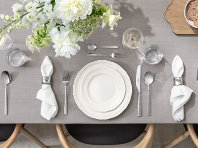 La table est dressée avec des assiettes blanches, des serviettes, des couverts, des verres et une composition florale sur une nappe grise.