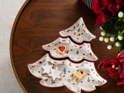 Dreiteiliges Toy's Delight Snackschalen-Set in Tannenbaum-Form Vogelperspektive auf ein dreiteiliges tannenbaumförmiges Porzellan-Snackschalen-Set der Kollektion Toy's Delight auf einem dunklen Holztisch, befüllt mit weihnachtlichen Zimtsternen und einem herzförmigen Keks, neben einem roten Geschenk mit grün-weiß gestreifter Schleife und roten Blumen.
