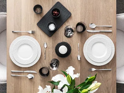 Elegant dining table with white Manufacture plates and Piemont cutlery Elegant dining table with white Manufacture plates, Piemont cutlery and decorative flowers on a wooden surface.
