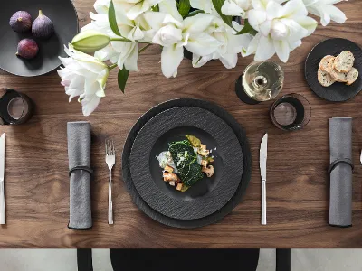 Manufacture by Villeroy & Boch for special occasions Elegant table setting with black Manufacture plates, cutlery, figs, a dish with vegetables and prawns, grey napkins, white flowers, a wine glass and sliced bread.