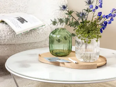 A round coffee table with a Villeroy & Boch Fleur vase holding blue and green flowers. In the background, an open magazine lies on a white, fluffy chair.
