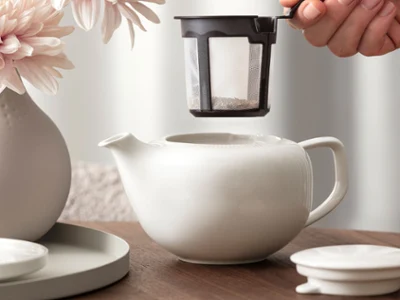 Tea Preparation with Afina A hand gently holds a tea filter over a white Afina teapot. The teapot stands on a wooden table, with a flower vase completing the scene in the background.