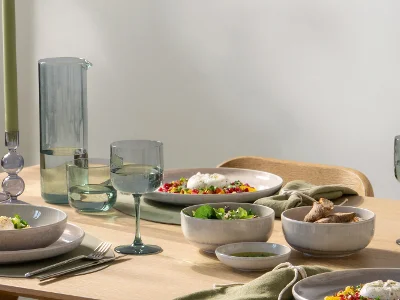 Like Carafe Table Set with Perlemor A modern dining table with a carafe, gray tableware, glasses, food, and green napkins in natural light.