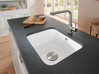 Lavello Cisterna con piano di lavoro antracite Un lavello sottopiano Cisterna bianco è incassato in un piano di lavoro color antracite. C'è una tavola di legno con sopra un coltello