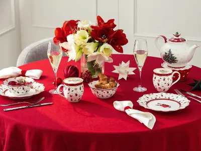 Colazione festiva di Natale con Toys Delight di Villeroy & Boch Tavolo rotondo con una tovaglia rossa apparecchiata per l'ora del tè, con porcellana festiva, pasticcini, fiori e decorazioni natalizie.