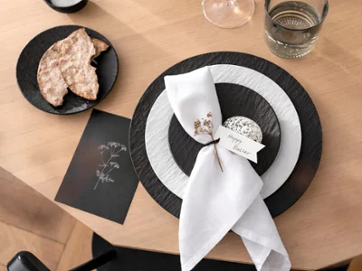 Elegant table setting with white and black plates, a wrapped napkin with a bow. Nearby are crispbreads on a small plate and a glass of water.