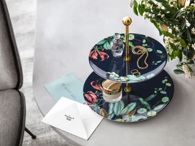 An Avarua tiered cake stand on a table with a small perfume bottle, jewellery and a jar of cream on top.