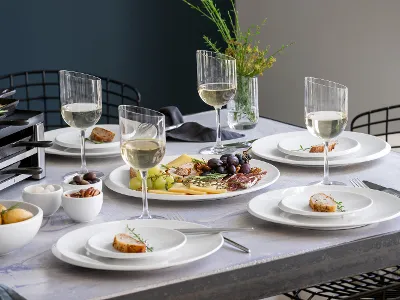 A dining table set with the elegant NewMoon collection from Villeroy & Boch, with four place settings, each served with a glass of white wine. In the center is a platter of cheese, grapes and figs, surrounded by bowls of potatoes, nuts, and olives.