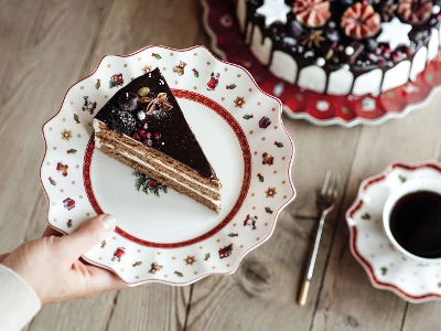 On the table are a decorated whole cake, a cup of coffee, a fork, and a saucer and plate set from Toy’s Delight. One hand holds a plate with a piece of chocolate cake. The table also features a decorated whole cake, a cup of coffee, a fork and a saucer, and plate set from Toy’s Delight.