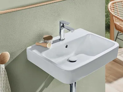 Lavabo O.novo bianco con rubinetteria cromata Un moderno lavabo rettangolare bianco O.novo con rubinetto cromato, spazzola e piccolo contenitore appoggiati accanto al rubinetto e davanti a una parete verde opaco. Sullo sfondo una sedia beige.