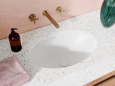 Loop & Friends vask i moderne badeværelse: Loop & Friends vask i moderne badeværelse Moderne vask med terrazzo bordplade, messinghane og sæbedispenser. Et lyserødt håndklæde ligger ved siden af, og i hjørnet står en vase med grønt glas.