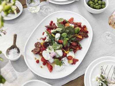 An Artesano plate from Villeroy & Boch with a mixed salad with tomatoes, herbs, and cold cuts on a white tablecloth. Next to it are bowls of olives, a glass of rosé, and a bowl of salt.