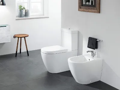 Inodoro Subway 2.0 junto a un bidé sobre un suelo de baldosas grises  Un moderno cuarto de baño con inodoro blanco y bidé sobre suelo de baldosas. A la izquierda hay un taburete de madera y un pequeño armario, en la pared hay una ventana y una obra de arte enmarcada.