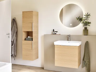 Architectura tap on a white washbasin with wooden furniture Modern bathroom with light-coloured wooden furniture, a wall-mounted washbasin and a round backlit mirror. Minimalist decor with a small plant on the shelf.