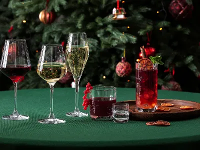 Bicchieri natalizi Toy’s Delight davanti a un albero di Natale decorato Un tavolo verde con cinque diversi bicchieri "Toy's Delight" di Villeroy & Boch, una tagliere con fette d'arancia essiccate e un albero di Natale decorato sullo sfondo.