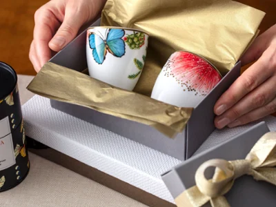 Hands holding an open gift box with two Amazonia cups Hands holding an open gift box with two Amazonia cups decorated with flower and butterfly motifs. A closed gift box with a bow stands nearby on a table.