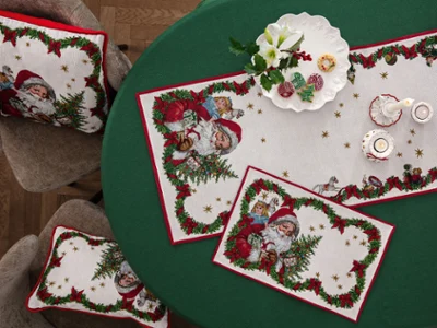 Toy's Fantasy Gobelin cushions and table textiles with Santa Claus motifs Top-down view of a green round dining table styled with Toy's Fantasy tapestry placemats, a runner, and matching cushions on chairs, decorated with a white porcelain plate of holiday cookies, candles, and a white Christmas rose.