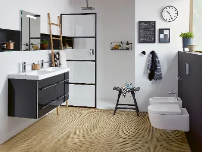 Bagno della collezione Avento di Villeroy & Boch Bagno moderno con lavabo doppio, doccia con cornice nera, pavimento in legno, WC e bidet a muro, un orologio, uno scaffale a scala e vari oggetti decorativi.