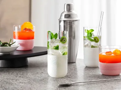 Four colorful cocktails in white Manufacture glasses on a marble surface, a cocktail shaker in the background, and a small bowl of bell peppers on a tray.