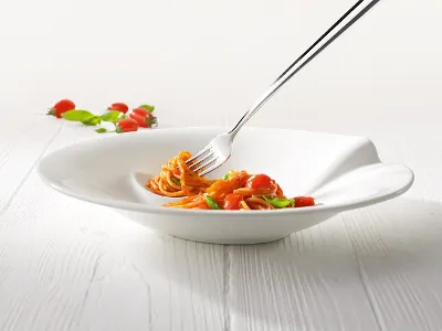 Pasta Passion spaghettibord met vork en pasta Een vork in een witte Villeroy & Boch Pasta Passion kom met een kleine portie spaghetti met kerstomaatjes en basilicum op een lichtgekleurde houten tafel.
