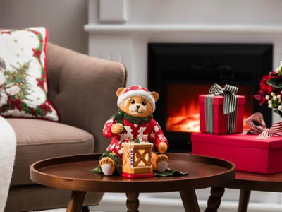 A teddy bear in pajamas, holding a lantern from Villeroy & Boch, sits on a coffee table next to gifts and flowers in a living room with a lit fireplace.