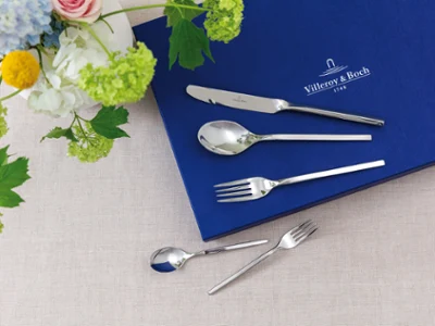 Bestekset met mes, lepel en vork op een blauwe doek naast een bloemstuk op een beige achtergrond.	