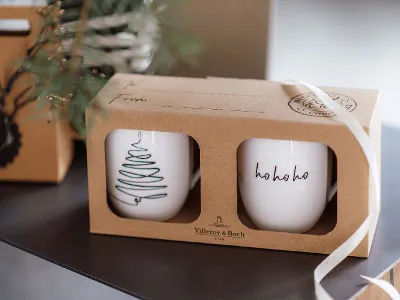 Two Statement cups in a cardboard box, one with a Christmas tree pattern and the other with the text "ho ho ho". The box is partly wrapped in a white bow.