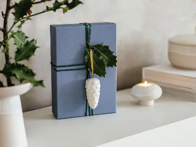Scatola regalo blu decorata con addobbo Winter Glow Scatola regalo blu, decorata con addobbo Winter Glow, accanto a una candela accesa e a libri su un tavolo.