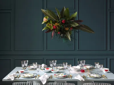 Stoviglie Amazonia su un elegante tavolo da pranzo di fronte a una parete con pannelli Il set da tavola Amazonia di Villeroy & Boch è composto da bicchieri, posate dorate e piatti con motivi floreali. Una composizione di fiori è appesa sopra il tavolo da pranzo, creando un bellissimo contrasto con la parete scura rivestita.