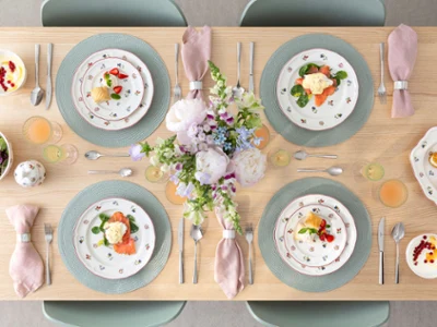 Petite Fleur, amorosamente dispuesta Una mesa de comedor preparada para cuatro personas con platos de flores Petite Fleur, servilletas rosas, bebidas y un arreglo floral.